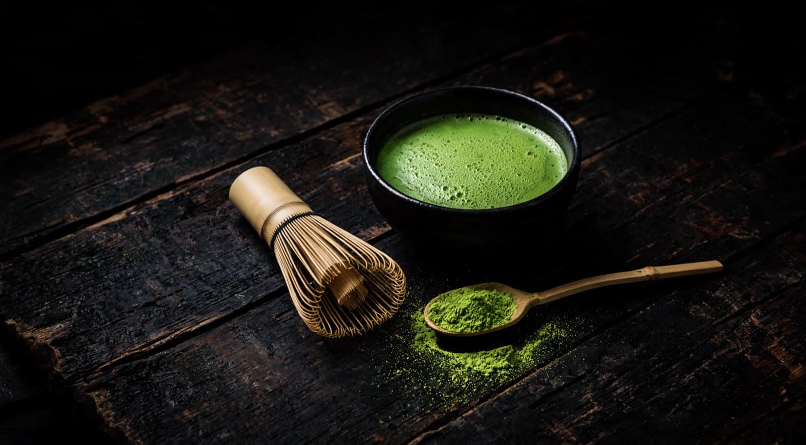 A dark ceramic chawan of freshly whisked first-harvest matcha, vivid jade under a fine even foam, beside a bamboo chasen and a chashaku holding dry sifted powder on weathered dark wood