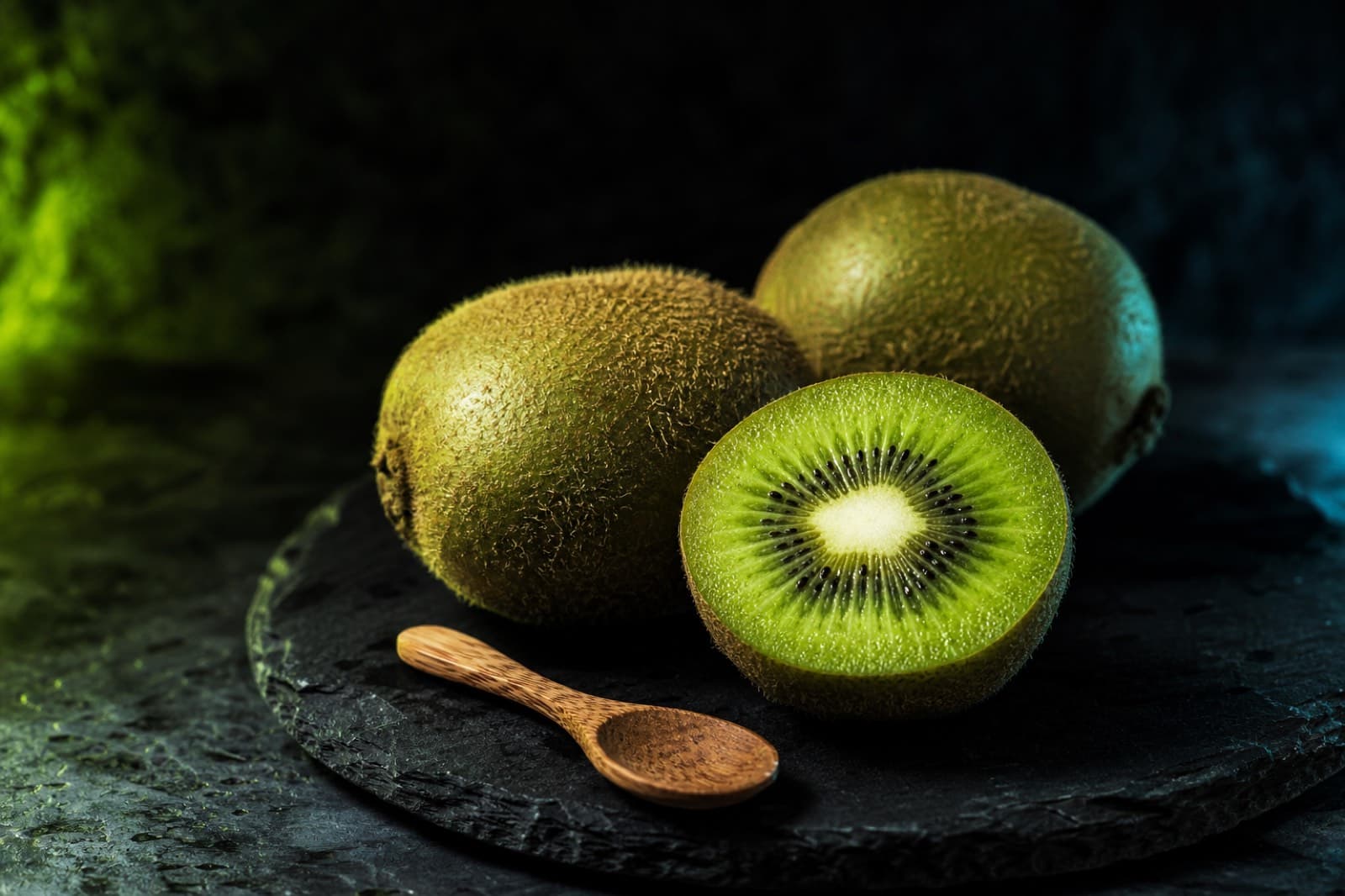 Whole and halved green kiwifruit with a wooden spoon on a dark slate surface