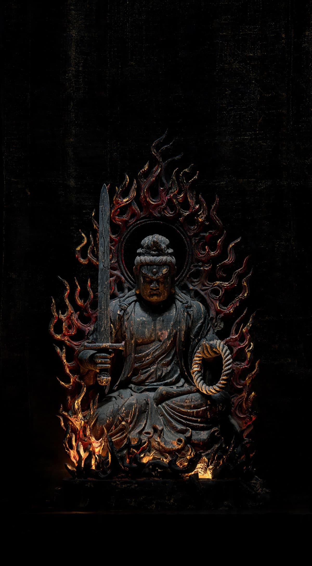 Painted woodcarving of Fudō Myōō seated wreathed in flame, sword in one hand and rope in the other, lit against deep black