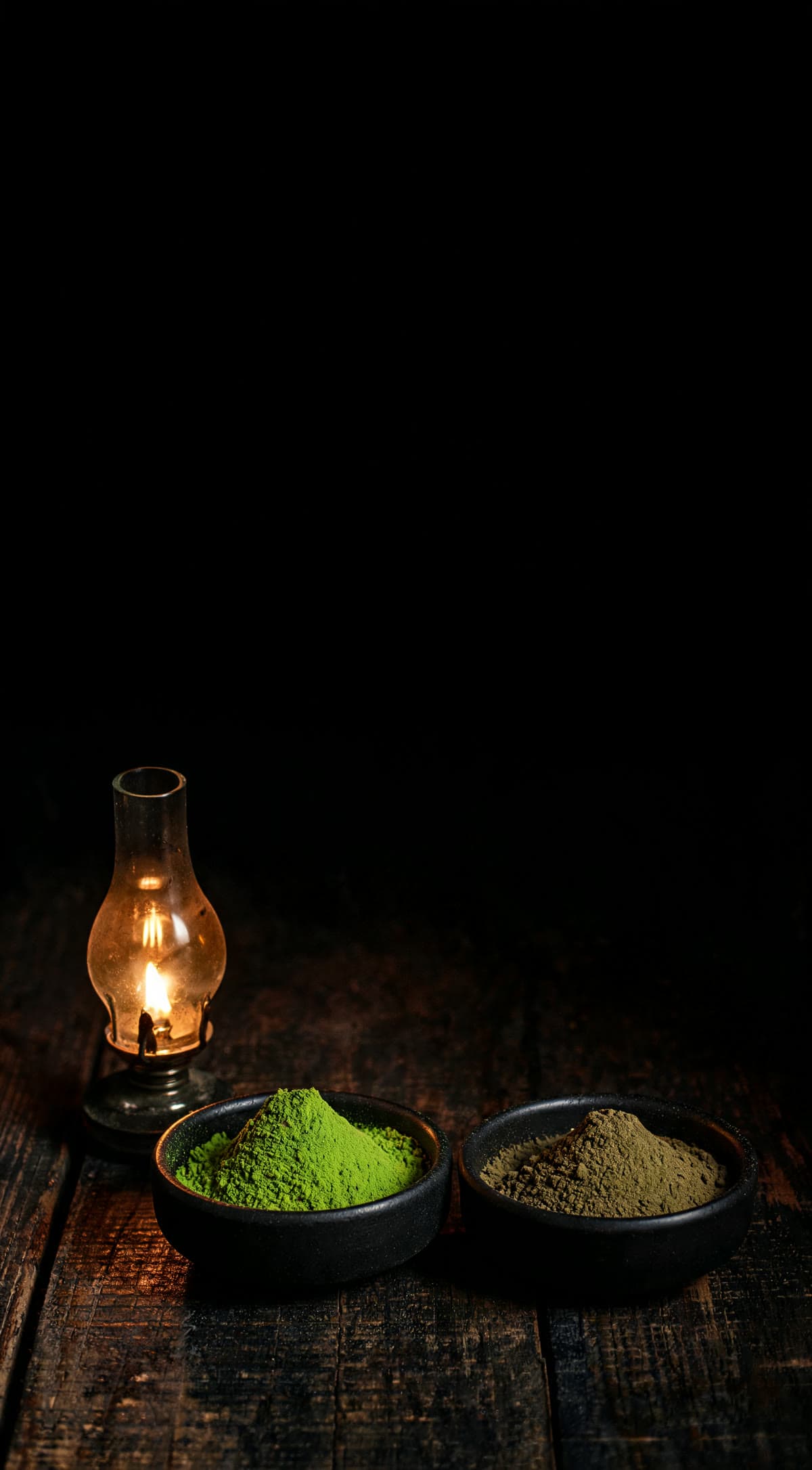 Two dark ceramic bowls of green tea powder lit by a single oil lamp — one vivid jade, one dull olive